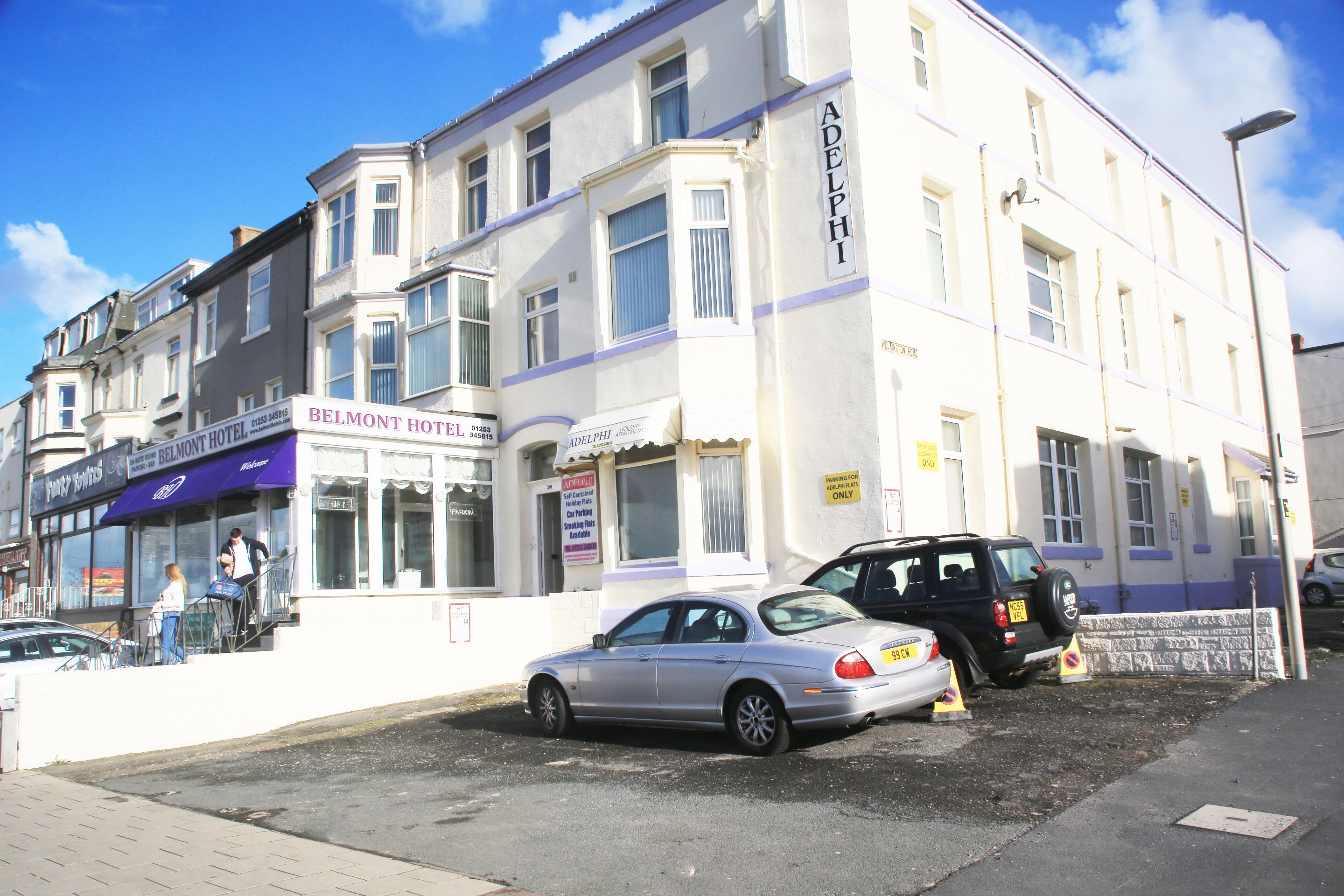 A hotel with a sign for 'BELMONT HOTEL' and 'ADELPHI' on the facade. There are cars parked in front, and a pedestrian is visible on the sidewalk.