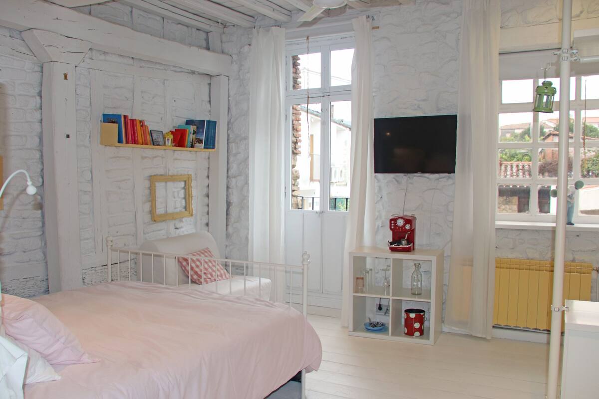 A bedroom with a bed, a white wardrobe, a shelf with books, a television, and a window with curtains.