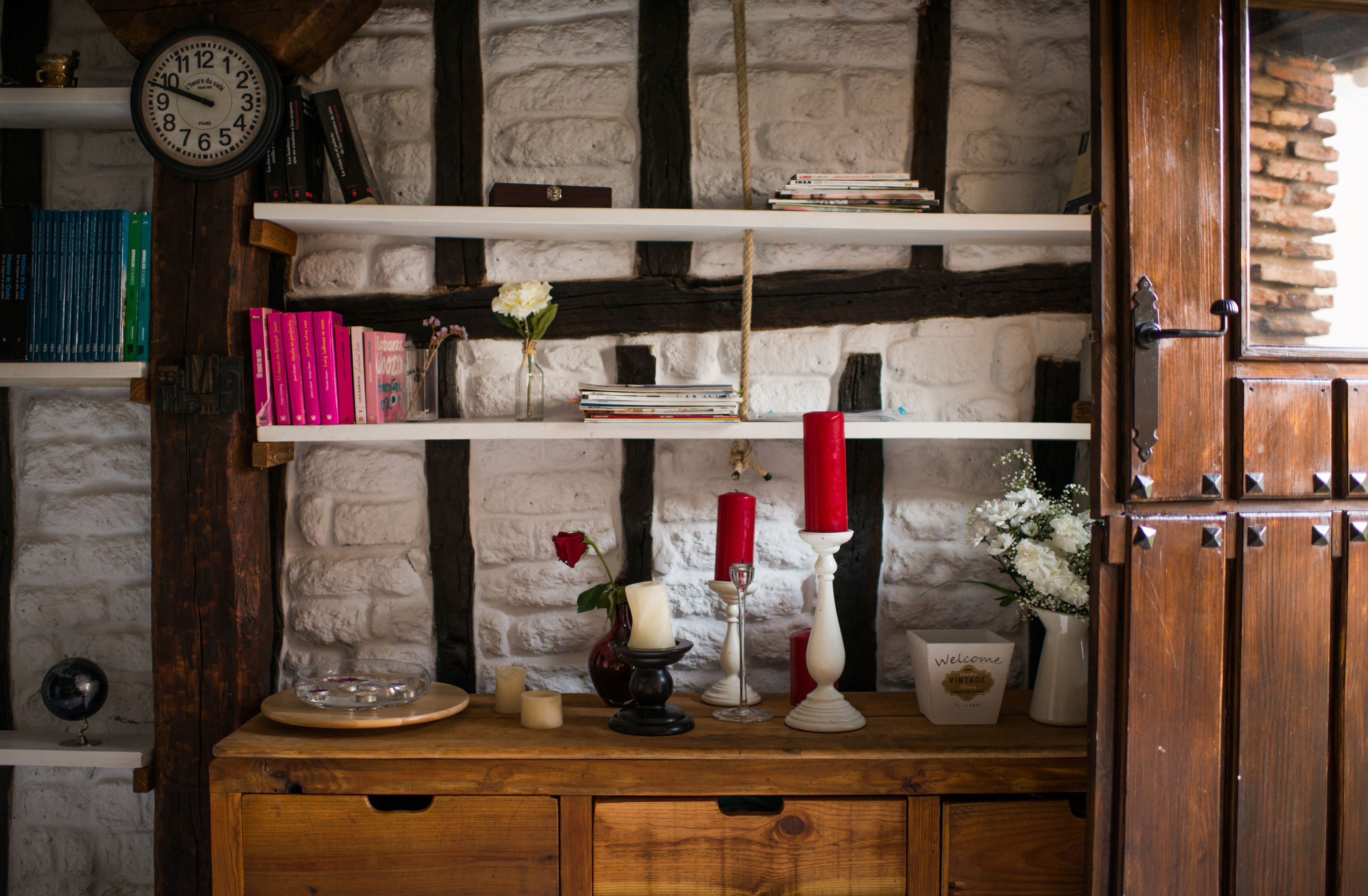 Una repisa de madera con libros, un reloj, velas y un florero con flores.