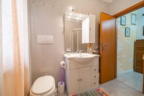 A bathroom with a toilet, sink, and mirror. There is a shower curtain, a toilet brush, and a rug on the floor.