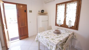 A room with a wooden door, a refrigerator, a table with a floral tablecloth, and a window with curtains. - Appartamento Le Conchiglie (Portoferraio)