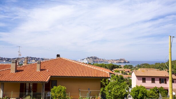 A residential area with terracotta-tiled roofs, a satellite dish, and a view of a distant town and coastline. - Appartamento Le Conchiglie (Portoferraio)