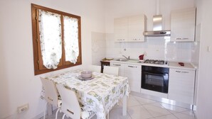 A compact kitchen with white cabinets, a gas stove, and a dishwasher. There is a dining table with chairs and a window with curtains. - Appartamento Le Conchiglie (Portoferraio)