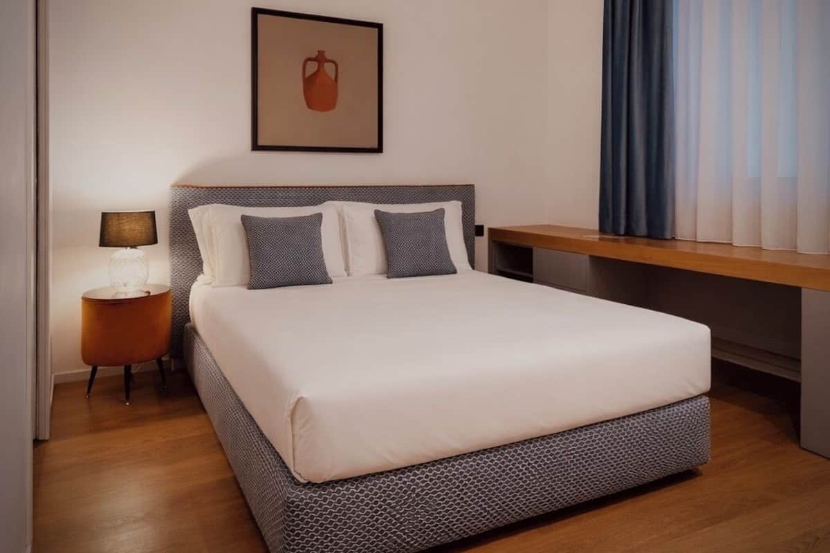A neatly arranged hotel room with a large bed, a bedside table with a lamp, a framed picture on the wall, and a window with curtains.
