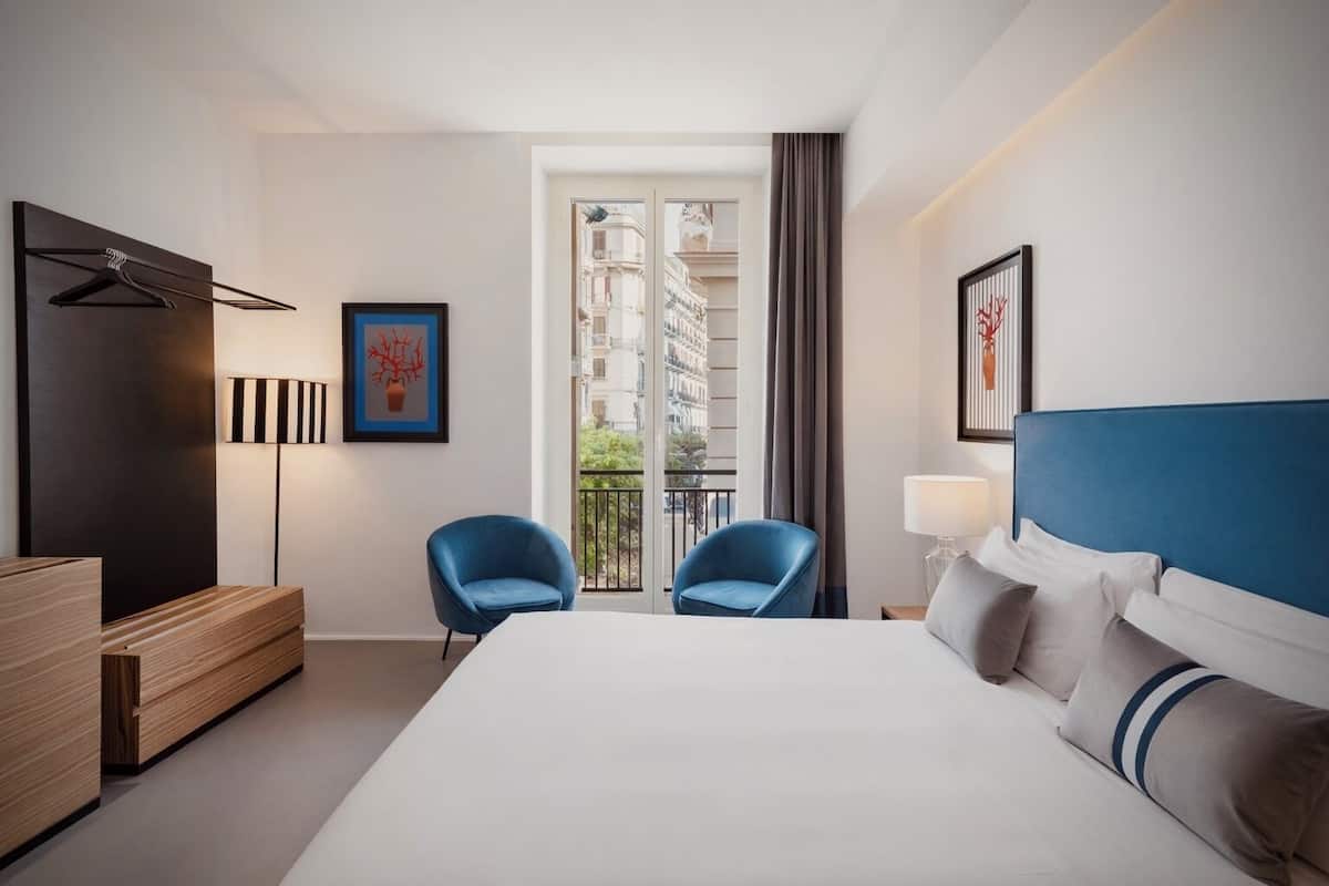 A modern hotel room with a large bed, two blue armchairs, a wooden dresser, and a view of the city through the balcony doors.