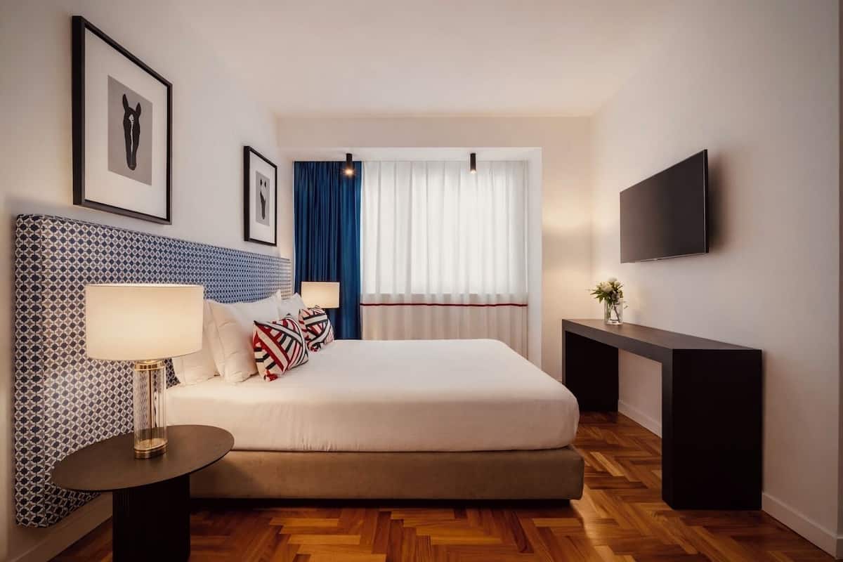 A modern hotel room with a large bed, a bedside table with a lamp, a TV mounted on the wall, and a window with curtains.