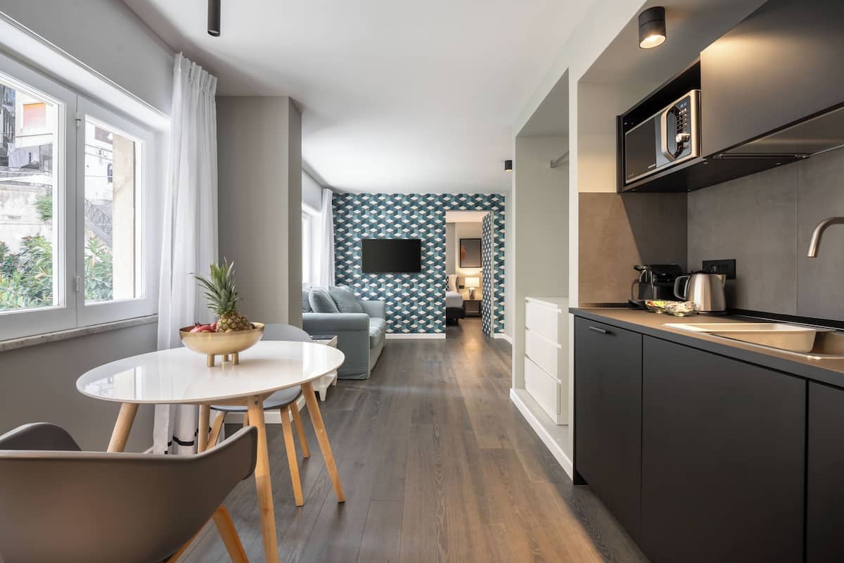 A modern kitchen with a built-in microwave, a coffee maker, and a sink. There is a dining area with a table and chairs, and a living room with a sofa and a TV.