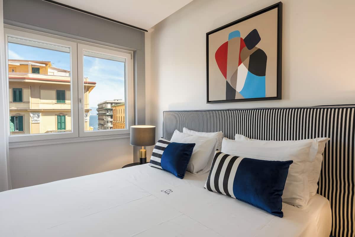 A hotel room with a bed, a window, a lamp, and a painting.