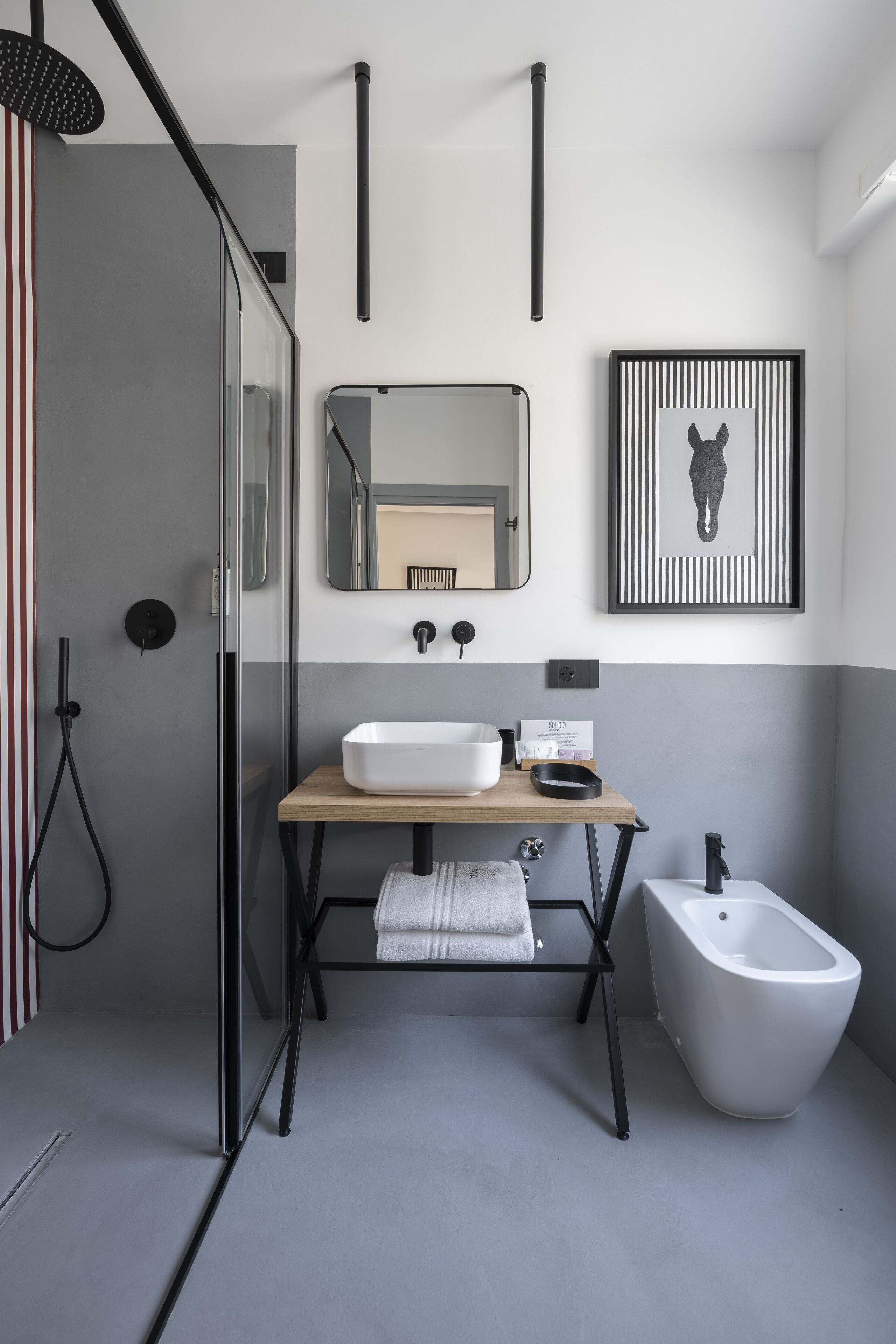 Un baño moderno con cabina de ducha de vidrio, un mueble de tocador de madera con lavamanos blanco, un bidé y una obra de arte enmarcada en la pared.