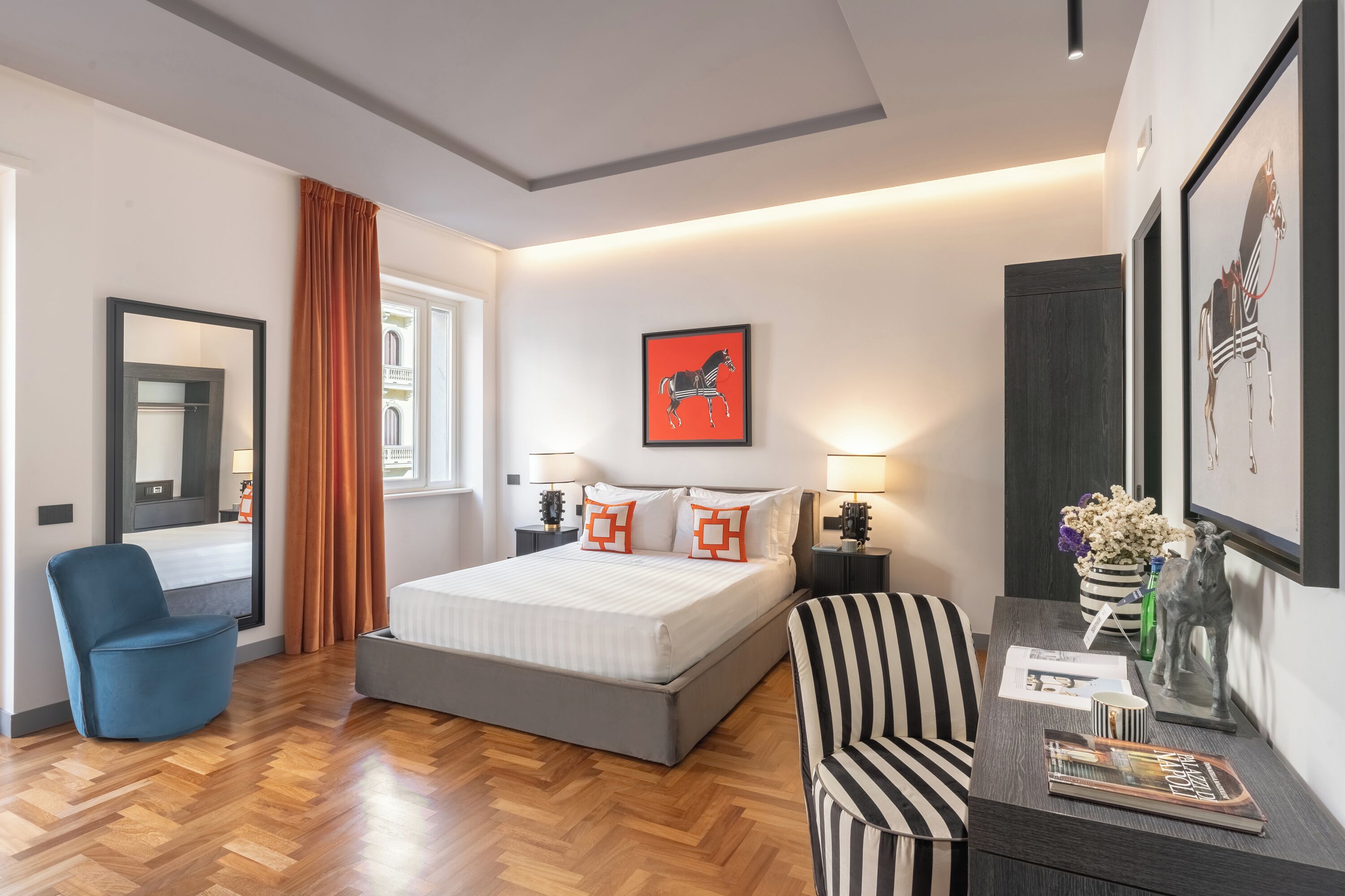 A modern hotel room with a large bed, a desk with a chair, a blue armchair, and artwork on the walls.