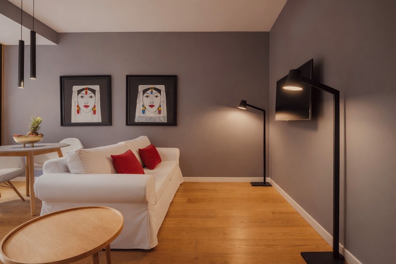 A modern living room with a white sofa, red cushions, a round wooden table, and two framed artworks on the wall.