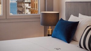 A hotel room with a bed, a window with a view of buildings and water, and a bedside lamp. - Le Mummarelle  (Naples)