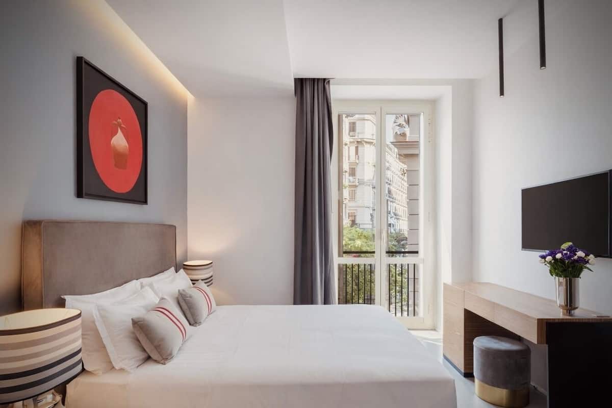 A modern hotel room with a large bed, a desk, a TV, and a balcony with a city view.