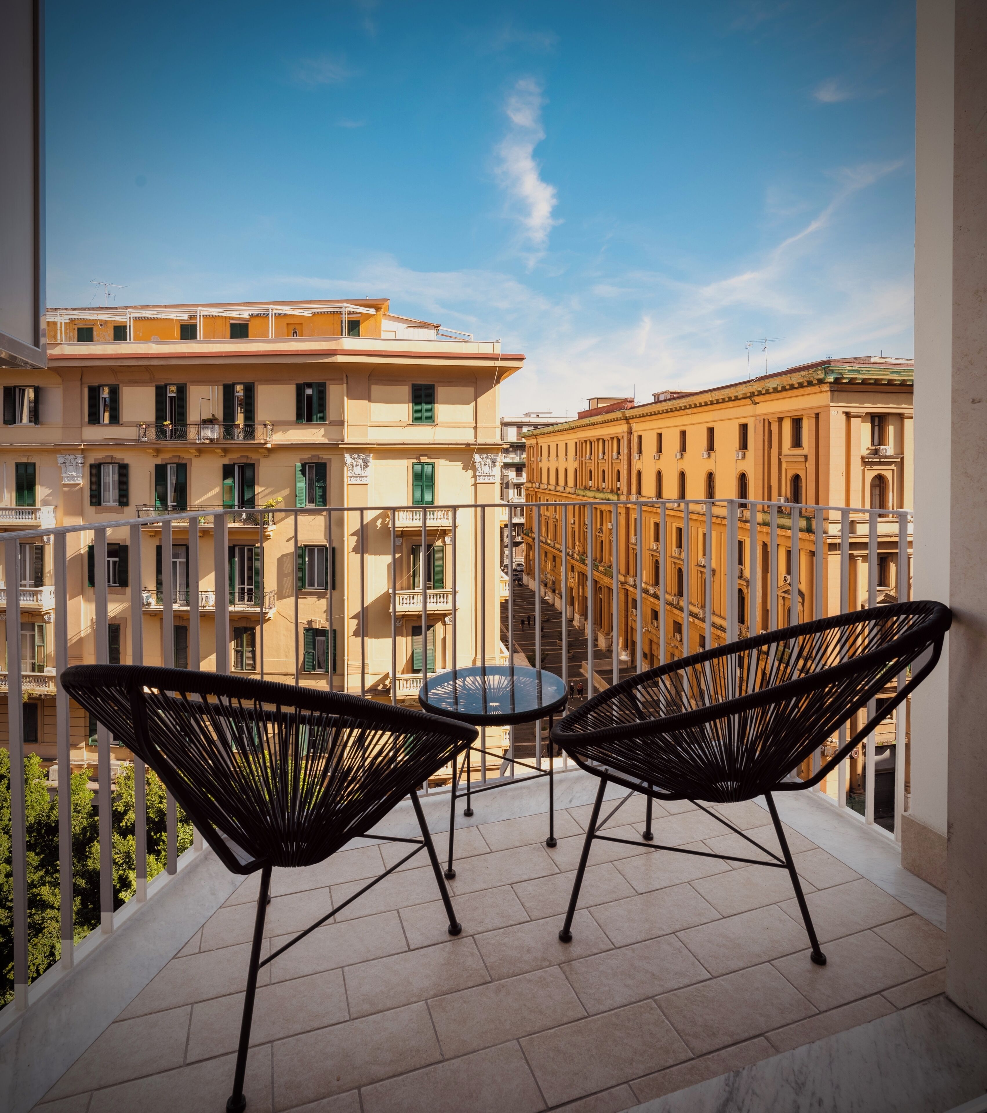 A balcony with two black chairs and a small table, overlooking a cityscape with multi-story buildings.