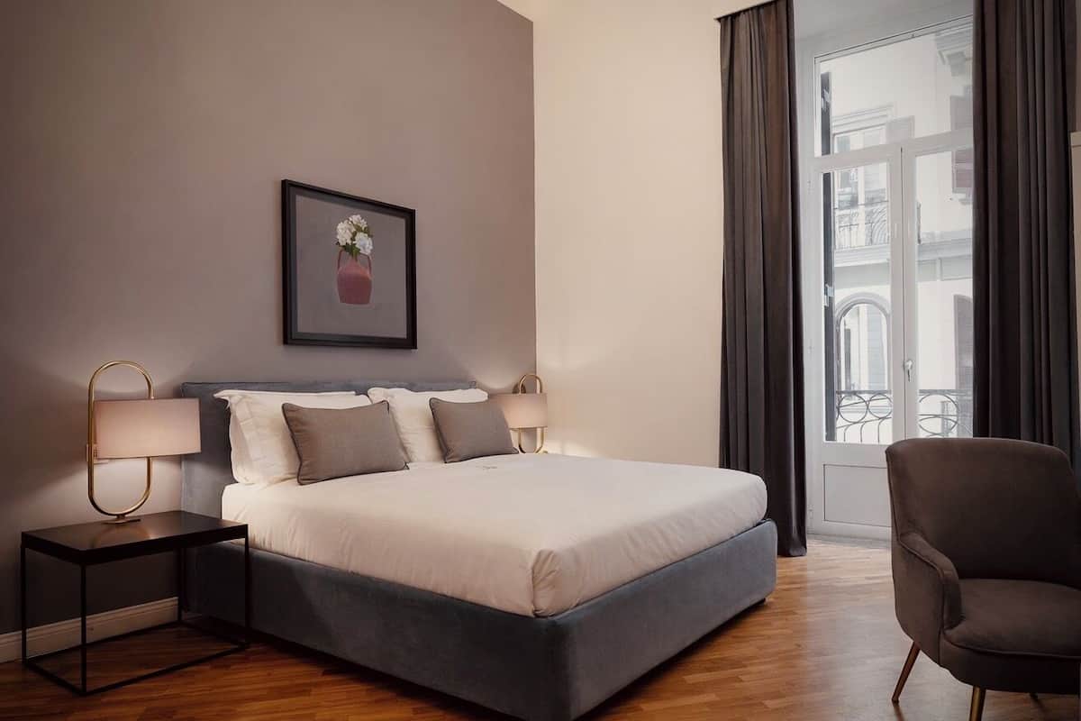 A modern bedroom with a large bed, two bedside tables with lamps, a chair, a framed picture on the wall, and a window with curtains.