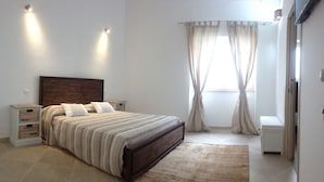 Una camera da letto con un letto, comodini, una testiera in legno, una finestra con tende e una piccola panca.