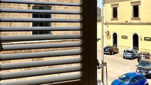 Una finestra con persiane, vista su un edificio con facciata in pietra e auto parcheggiate all'esterno.
