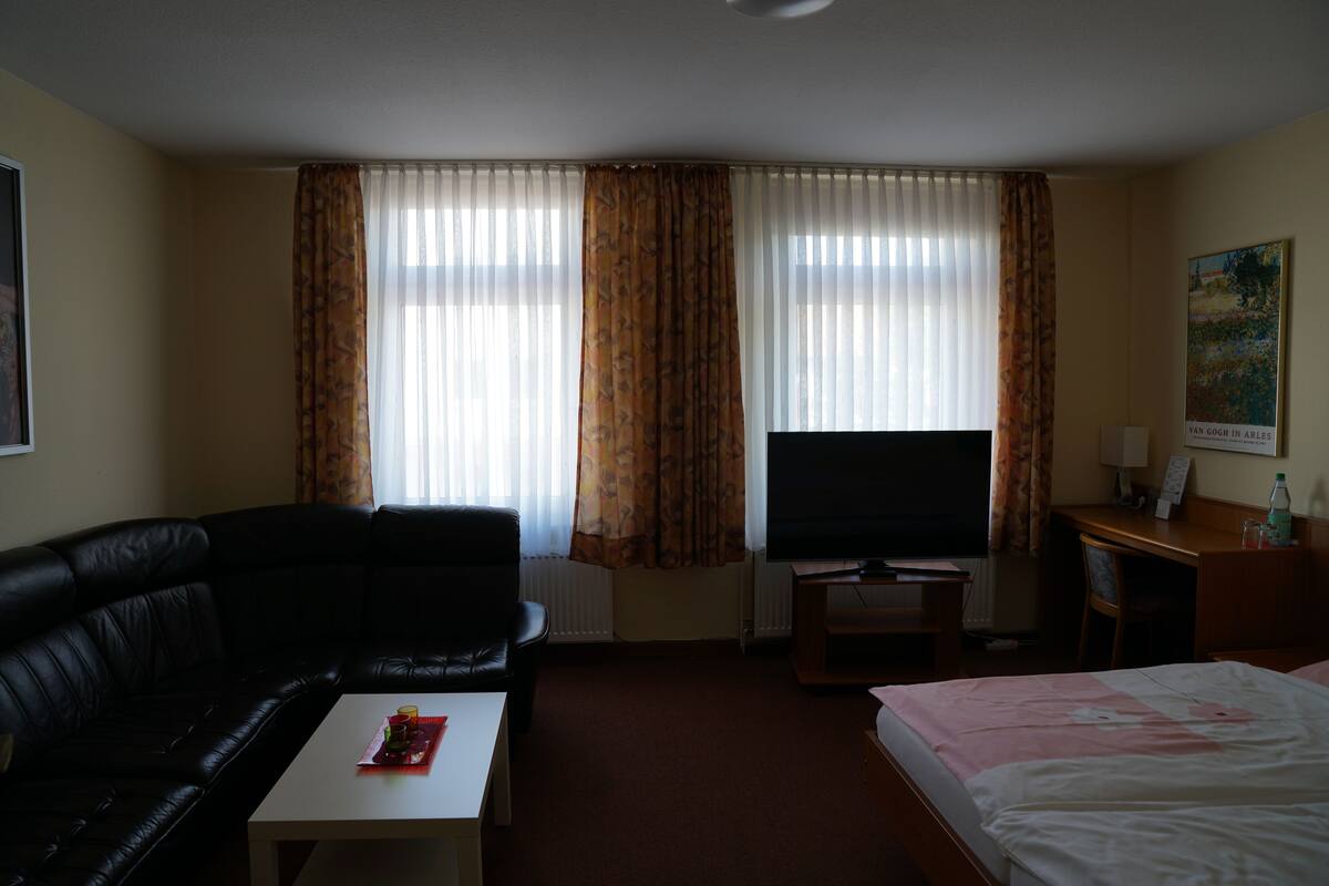 Une chambre d’hôtel avec un canapé en cuir noir, un téléviseur posé sur un meuble, une petite table avec un plateau, un lit recouvert d’une couverture rose et un tableau accroché au mur.