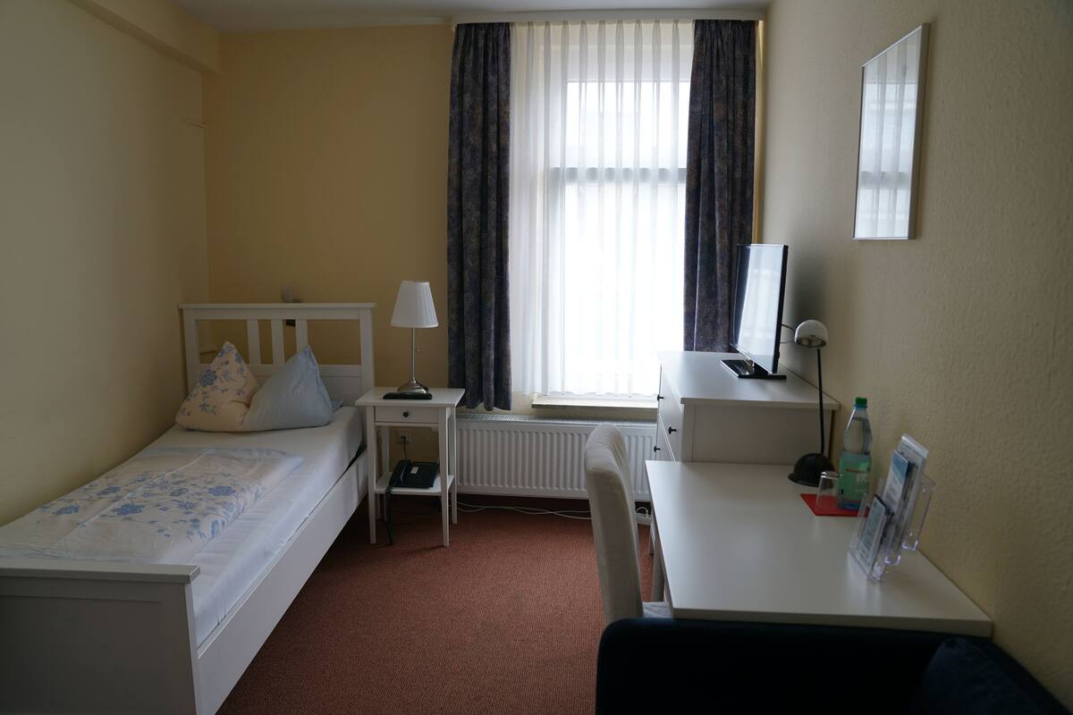 Une chambre d’hôtel avec un lit simple, un bureau, une chaise, un radiateur et une fenêtre avec des rideaux.