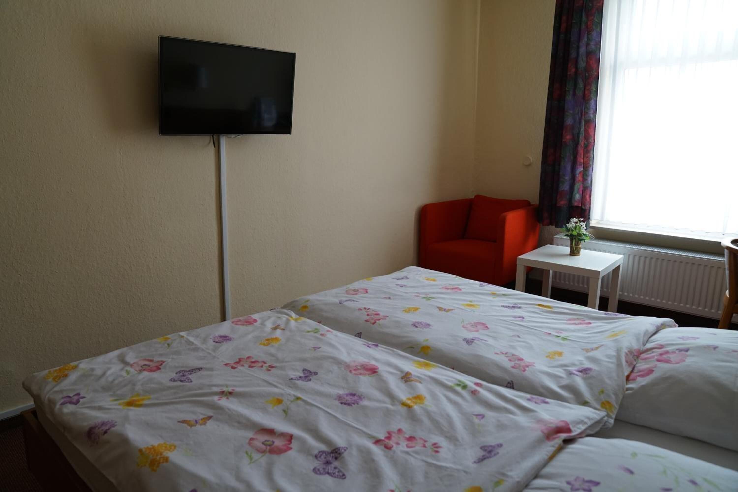 Habitación de hotel con dos camas, un televisor montado en la pared, una silla roja, una mesita con un florero y una ventana con cortinas.