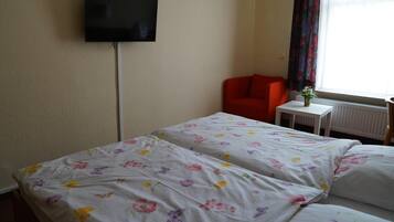 Habitación de hotel con dos camas, un televisor montado en la pared, una silla roja, una mesita con un florero y una ventana con cortinas.