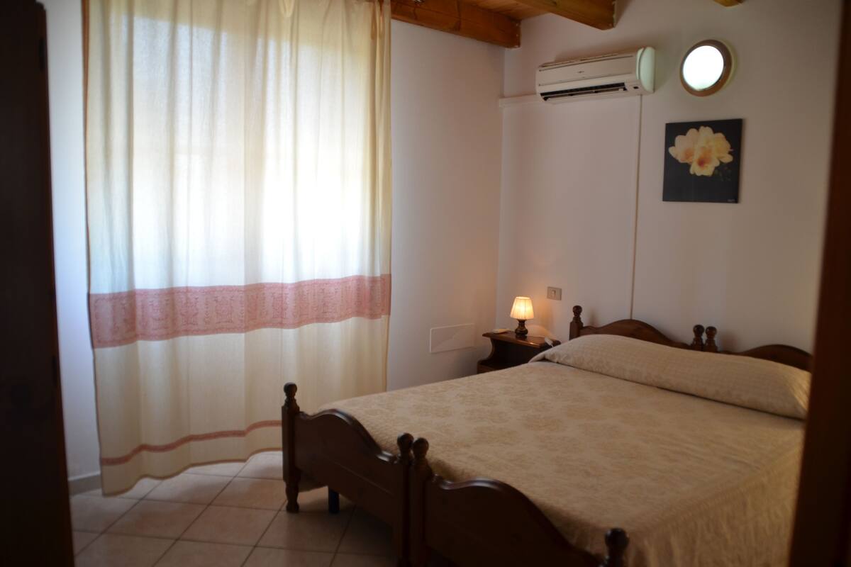 Un dormitorio con cama, mesita de noche, lámpara, equipo de aire acondicionado, ventana con cortinas e imagen colgada en la pared.