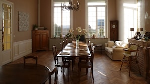 A dining room with a large wooden table, chairs, and a chandelier. There is a sofa and a clock on the wall.