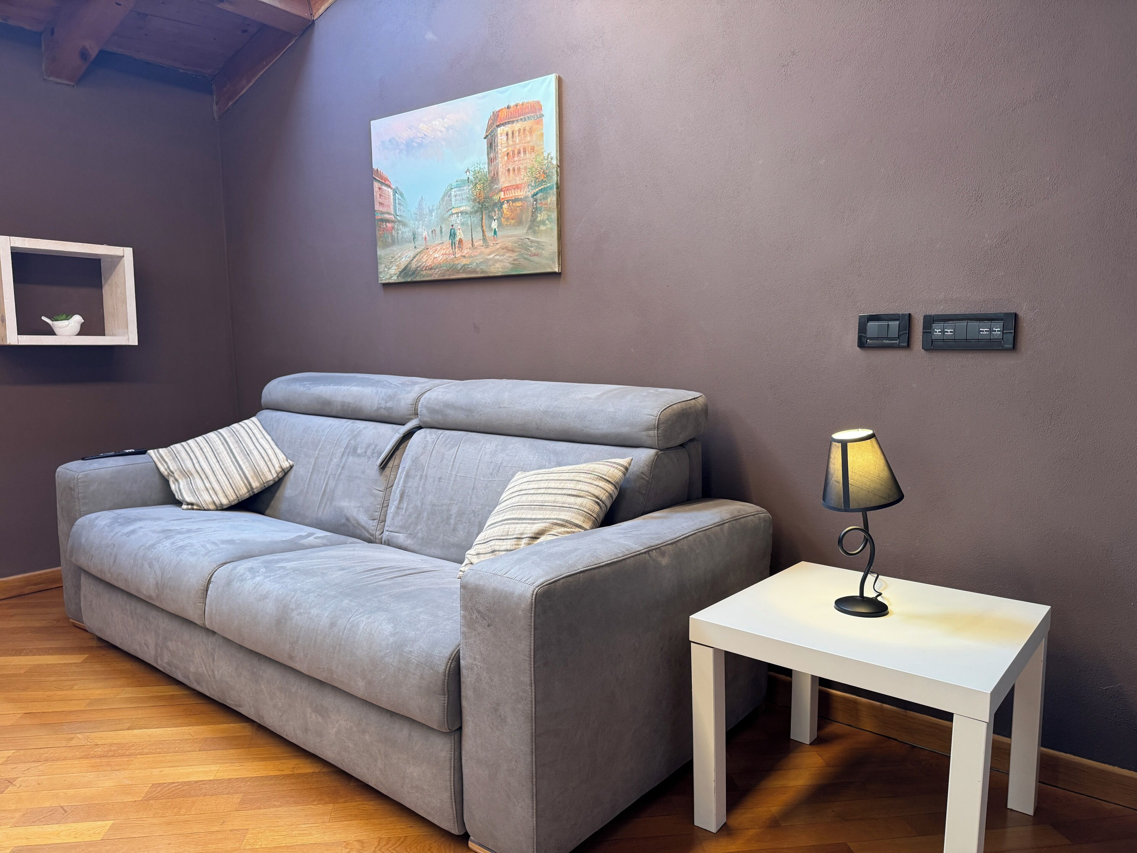 A grey sofa with striped pillows, a white side table with a lamp, and a painting of a street scene on the wall.