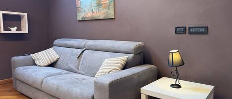A grey sofa with striped pillows, a white side table with a lamp, and a painting of a street scene on the wall.