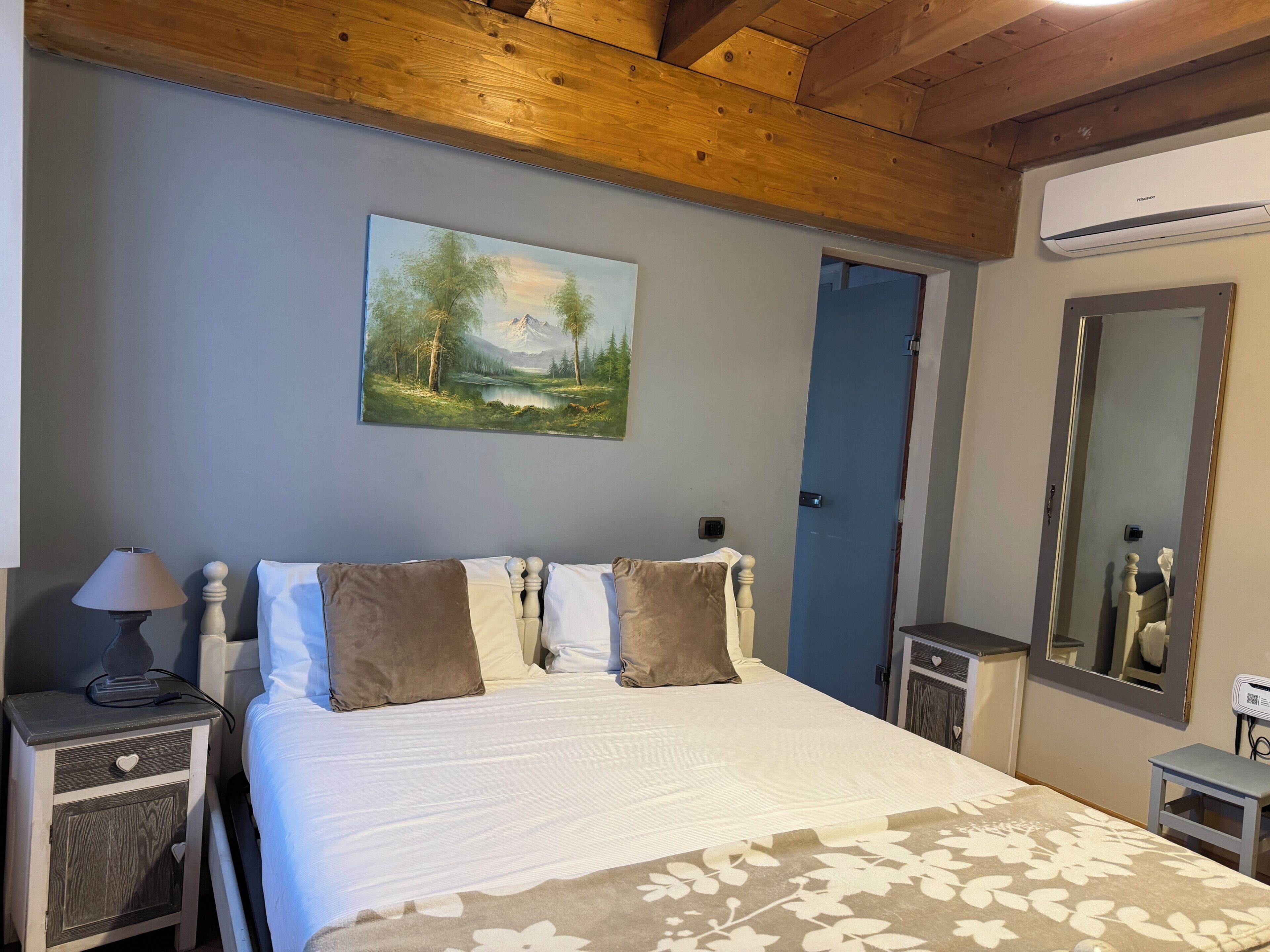 A bedroom with a bed, a nightstand, a lamp, a painting, a mirror, and an air conditioning unit.