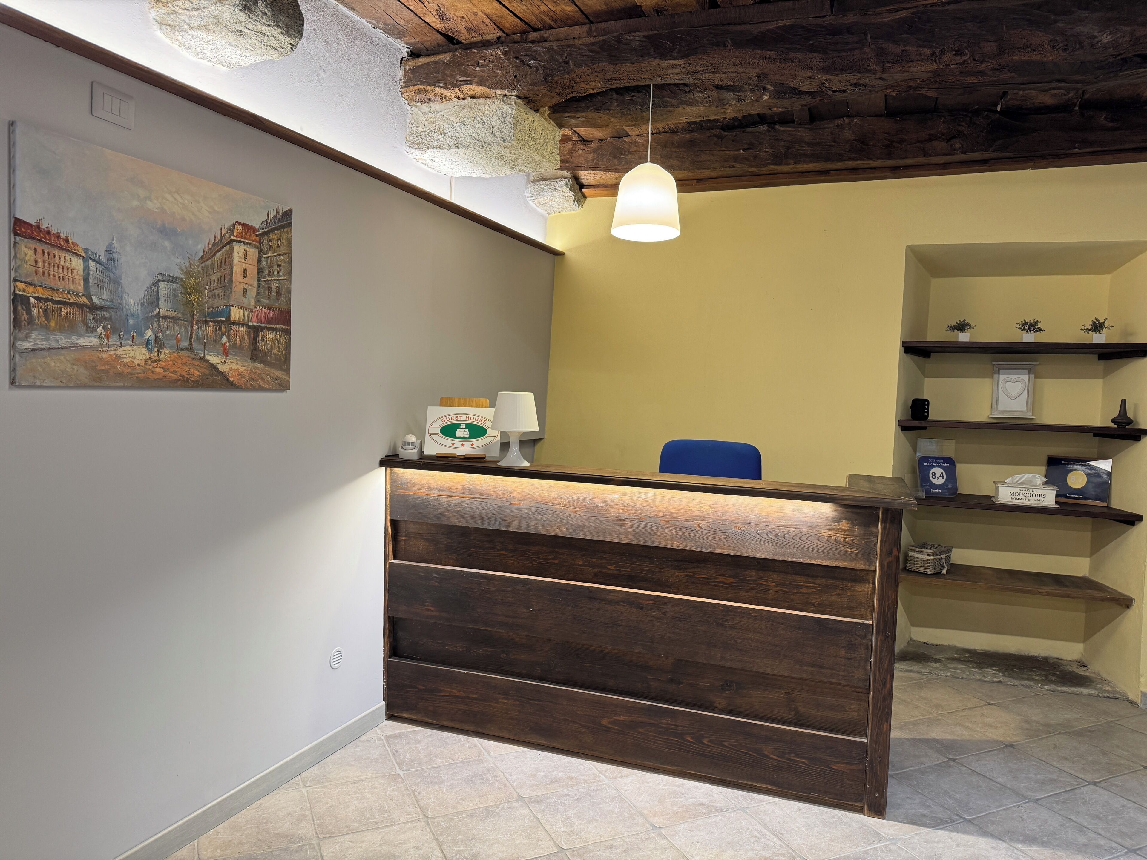 Un comptoir d’accueil avec un revêtement en bois, un luminaire suspendu, un tableau représentant une scène de rue et une étagère contenant divers objets, notamment une horloge et une boîte étiquetée « MOUCHOIRS ».