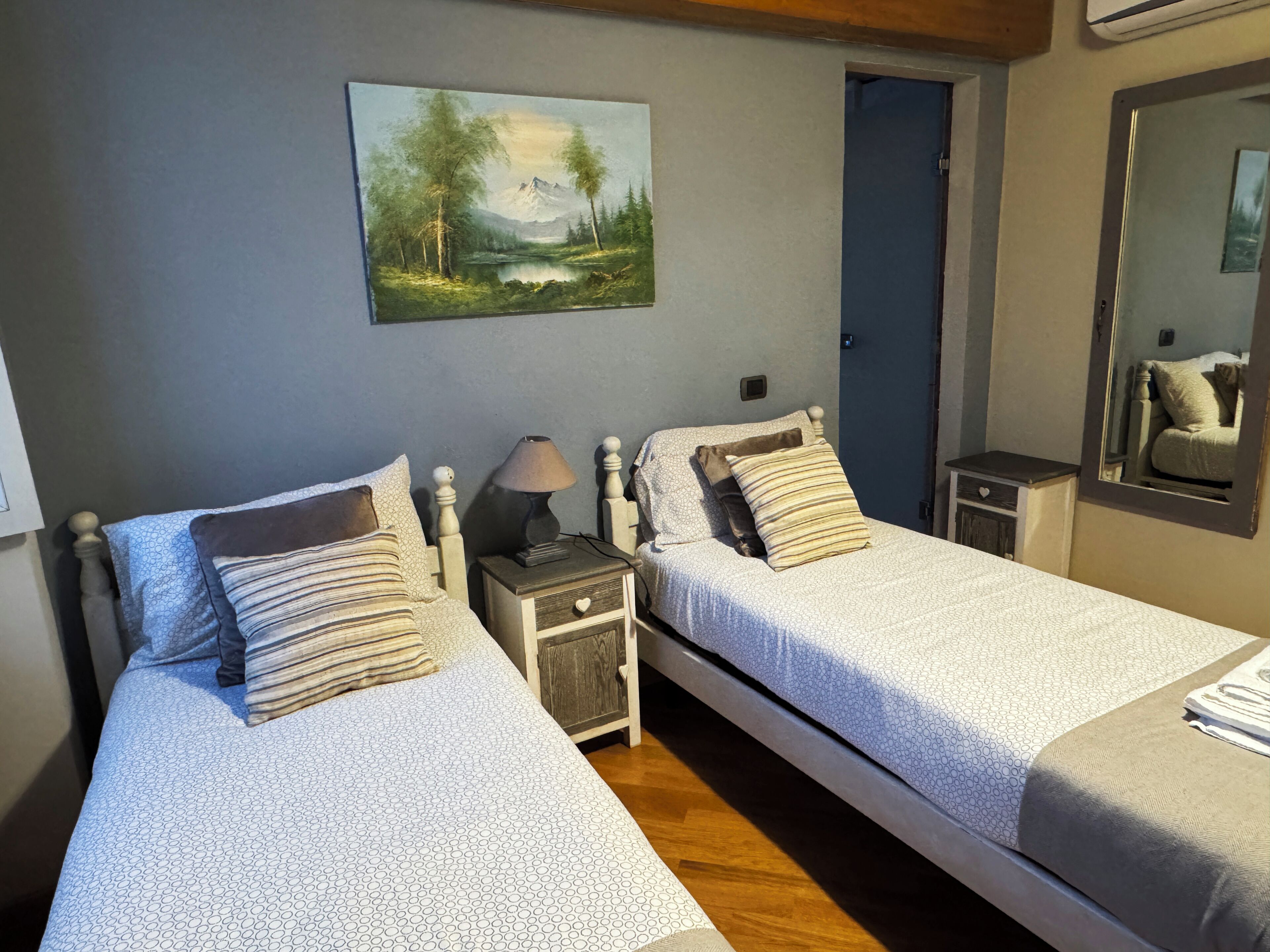 A bedroom with two beds, a nightstand with a lamp, a painting on the wall, and a mirror.