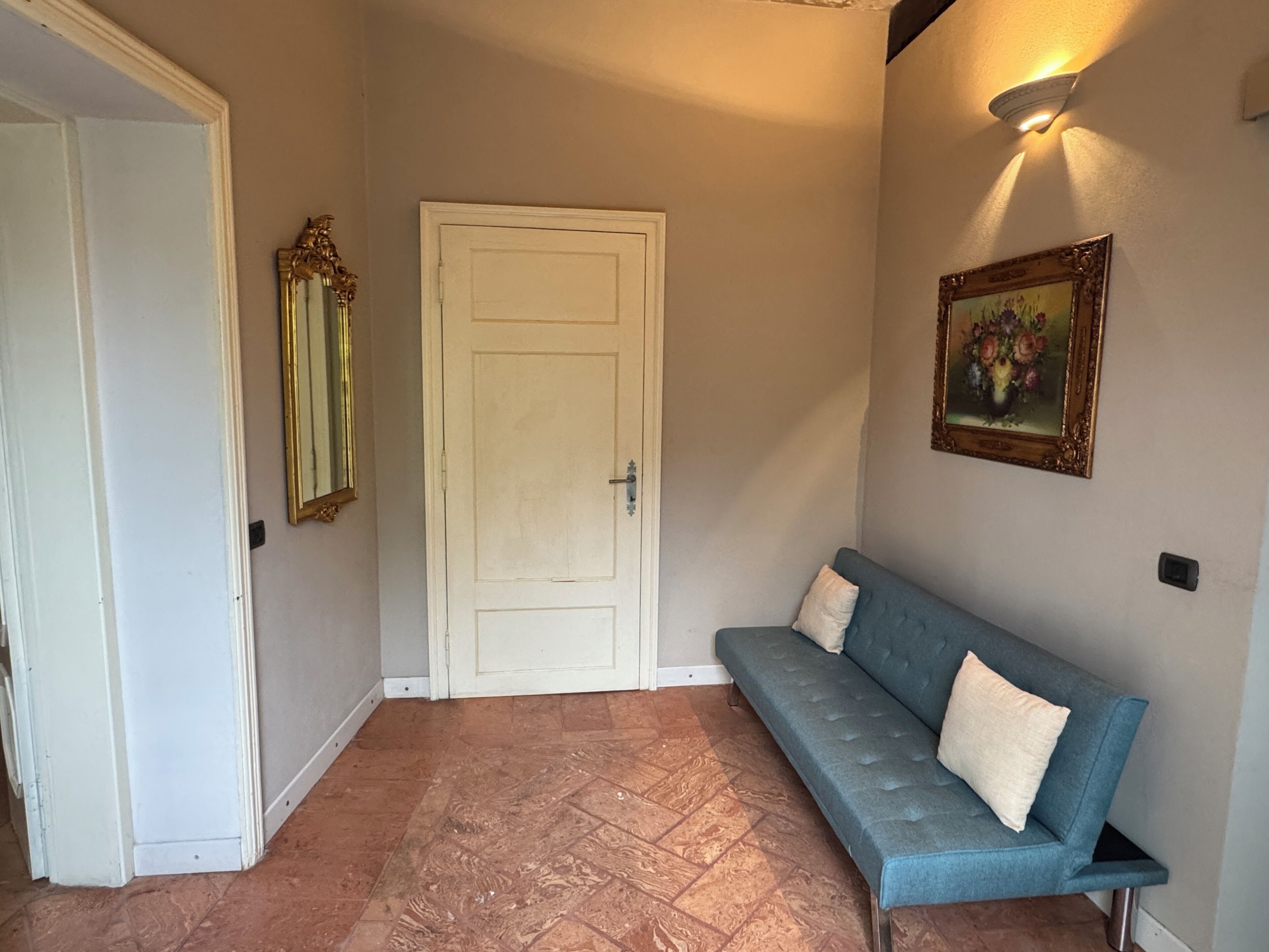 Un pasillo con una puerta blanca, un sofá azul con cojines, un espejo fijado a la pared y un cuadro enmarcado.