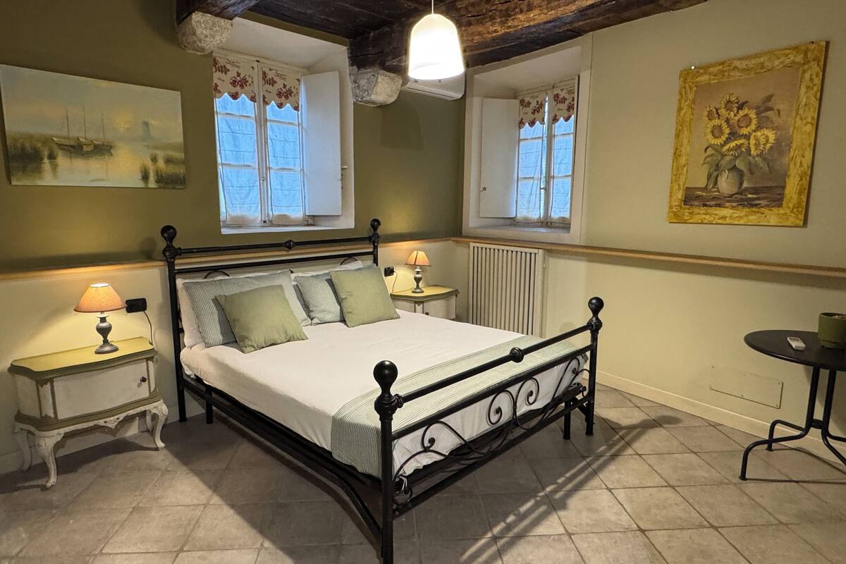 A bedroom with a bed, two bedside tables, a radiator, a window with a view, and a painting on the wall.