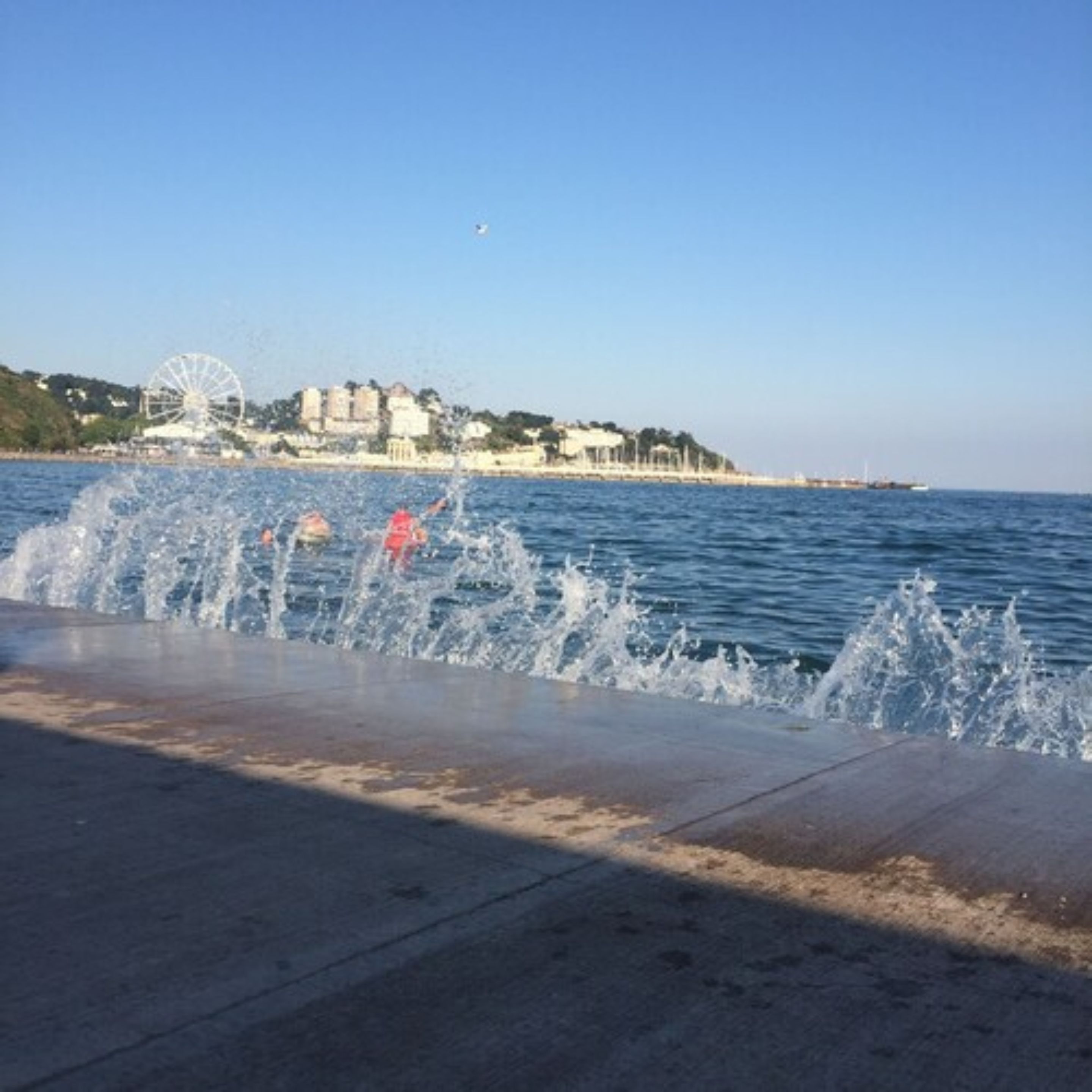 A waterfront promenade with water jets, a clear sky, and buildings in the background.