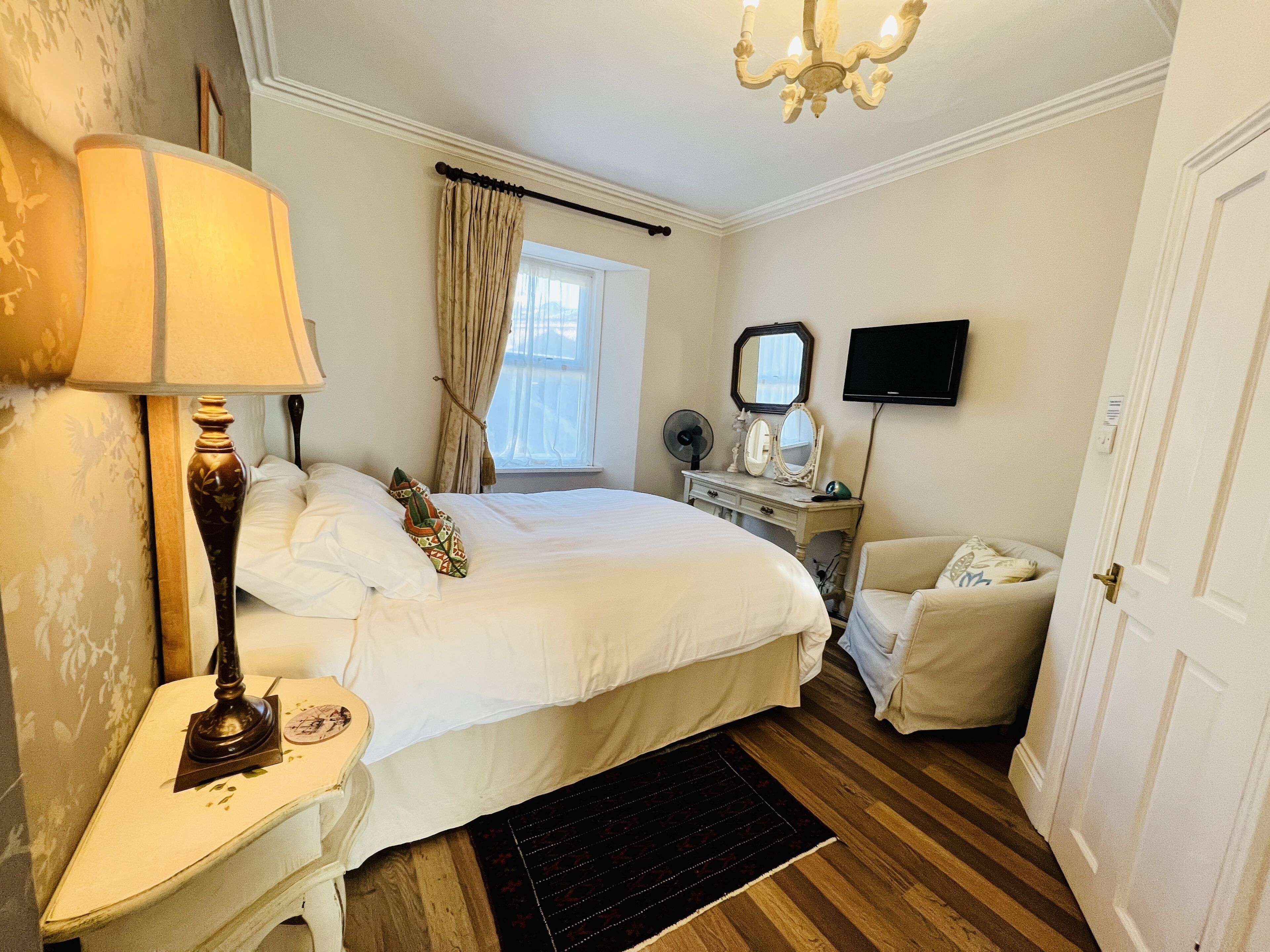 A hotel room with a large bed, a bedside table with a lamp, a dresser with a mirror, a television on the wall, and a door.