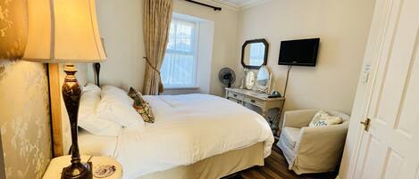 A hotel room with a large bed, a bedside table with a lamp, a dresser with a mirror, a television on the wall, and a door.