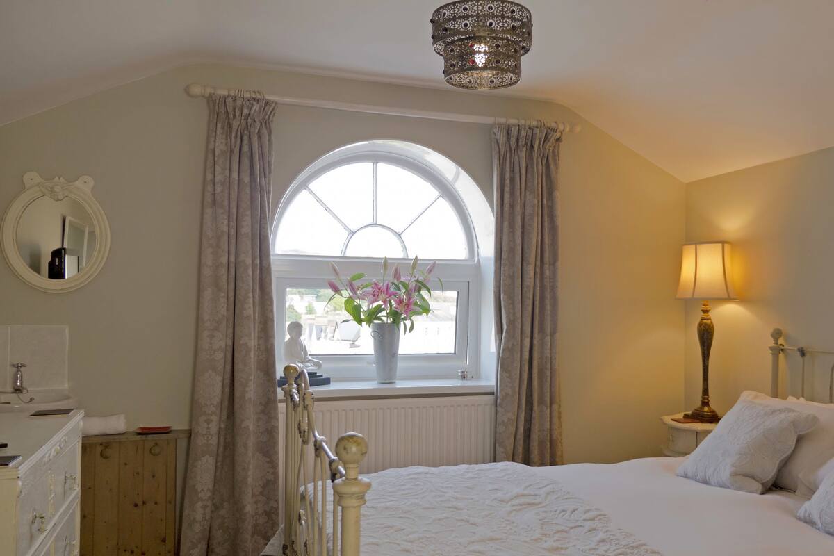 A bedroom with a large bed, a bedside table with a lamp, a window with curtains, a mirror, and a radiator.