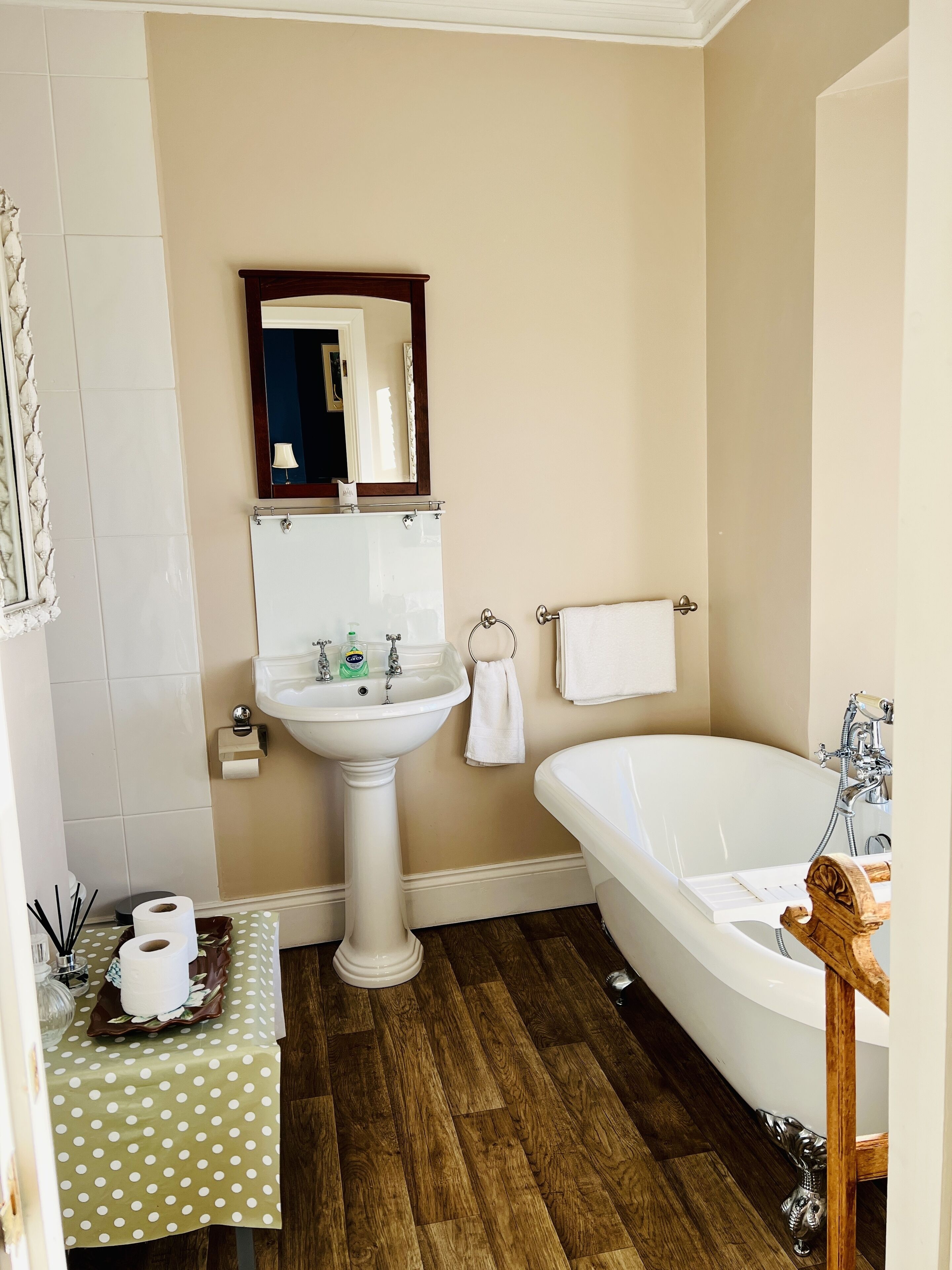 A bathroom with a pedestal sink, a large mirror, a bathtub, and a wooden floor.