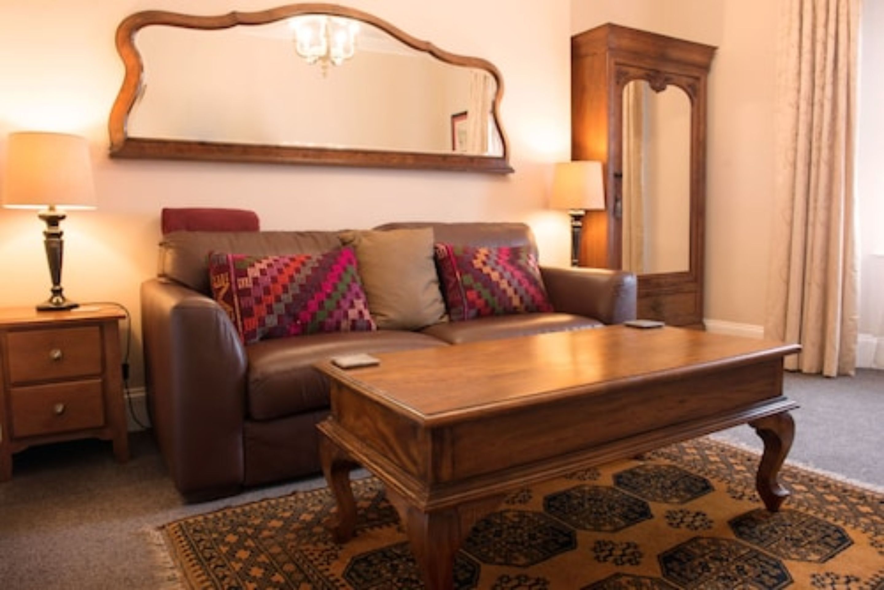 Una sala de estar con un sofá de cuero marrón, una mesa de centro de madera, un tapete estampado, una mesita auxiliar de madera con una lámpara y un espejo grande en la pared.
