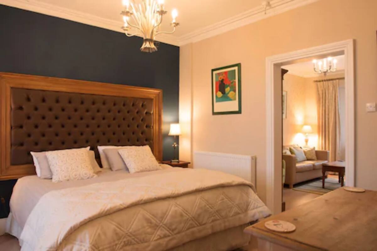 A hotel room with a large tufted headboard, a bed with a quilted cover, a bedside table with a lamp, a painting on the wall, and a view into an adjacent room with a sofa and a lamp.