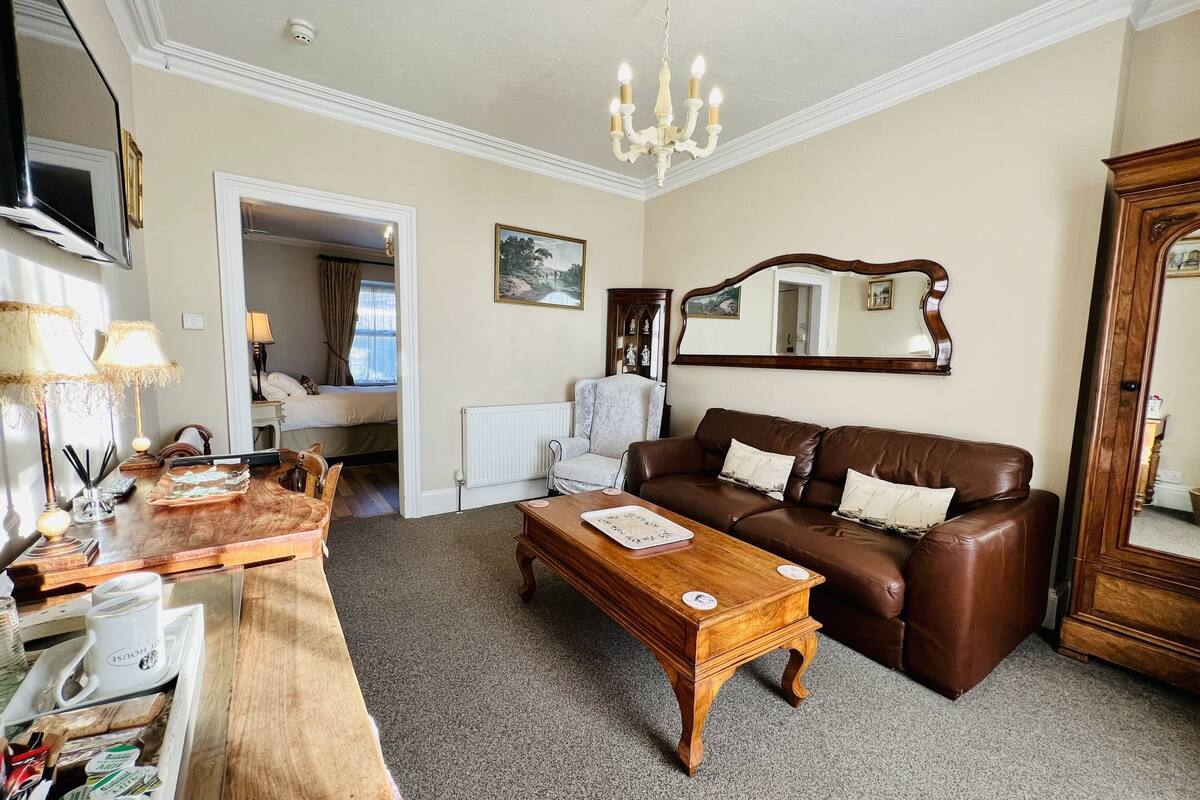 A cozy living room with a brown leather sofa, a wooden coffee table, and a dining area with a tray of coffee cups. There is a white radiator, a chair, and a mirror on the wall.