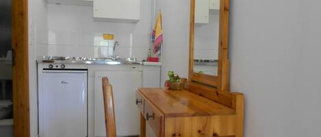 A compact kitchen with white cabinets, a sink, and a small refrigerator. There is a wooden table with chairs and a mirror on the wall.