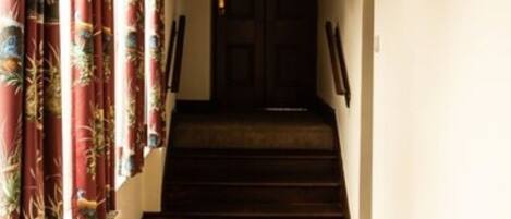 A carpeted hallway with a dark wooden staircase, white walls, and a door at the end.