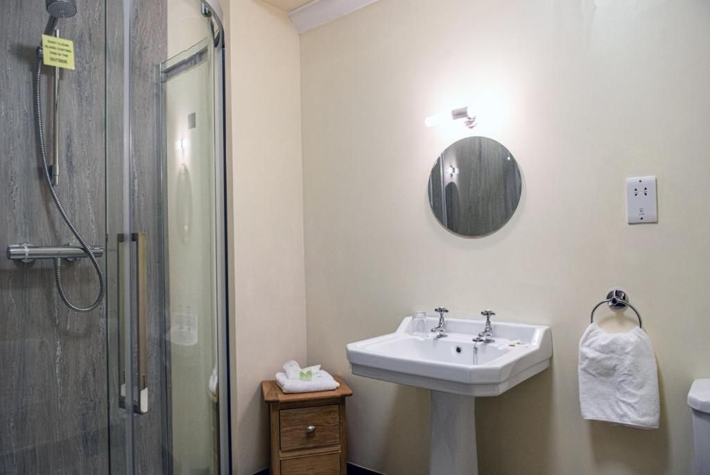 A bathroom with a glass-enclosed shower, a white sink, and a small wooden nightstand with a towel.