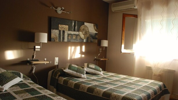 A bedroom with two beds, a wall-mounted lamp, a painting, and a window with curtains. - Hostal Delgado (Benabarre)