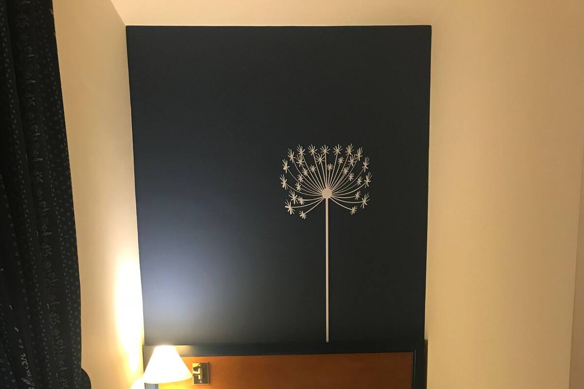 A hotel room with a bed, a nightstand with a lamp, a wall-mounted artwork, and a patterned rug.