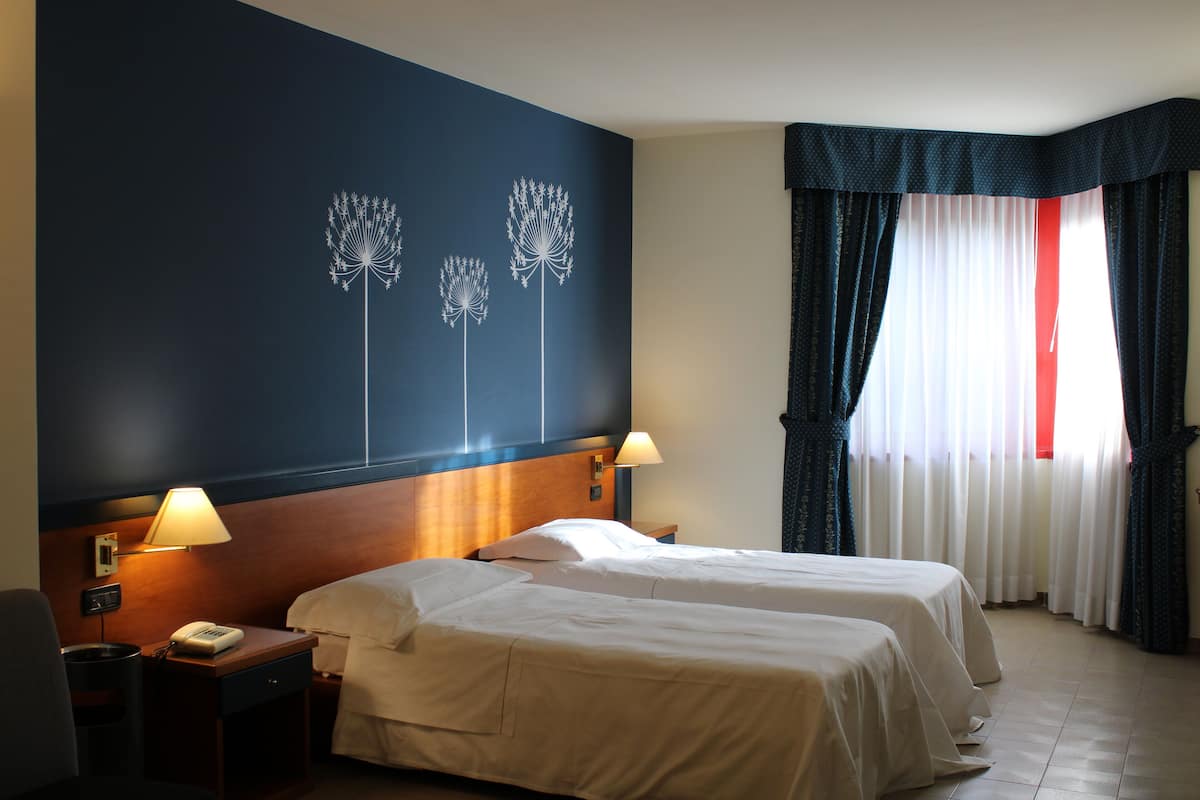 A hotel room with two beds, a nightstand with a lamp, a wall with a dandelion design, and a window with curtains.