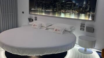 Un dormitorio moderno con una cama redonda, vistas a la ciudad y una mesita de noche redonda iluminada.