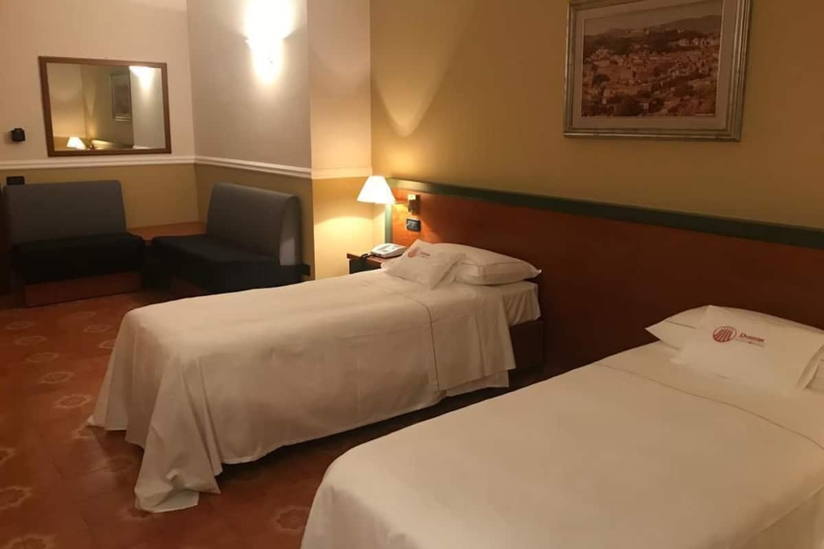 A hotel room with two single beds, a desk with a lamp, a chair, a nightstand with a phone, and a framed picture on the wall.